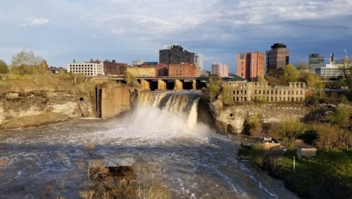 A waterfall cascades over rocks, with a city skyline in the background under a partly cloudy sky.