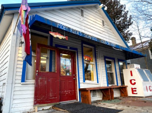 A quaint seafood shop with a red door and colorful flags, featuring an "Open" sign and an ice display outside.