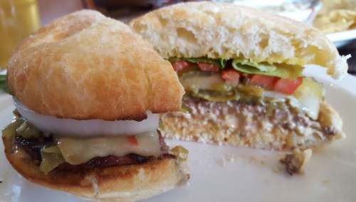 A close-up of a sandwich with a toasted bun, layers of beef, cheese, lettuce, tomato, pickles, and onions.