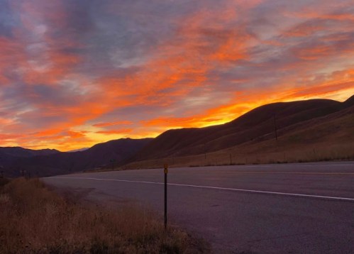 A vibrant sunset paints the sky in orange and purple hues over a quiet road and rolling hills.