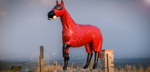 A large red horse statue wearing a mask stands on a hill with a city skyline in the background.