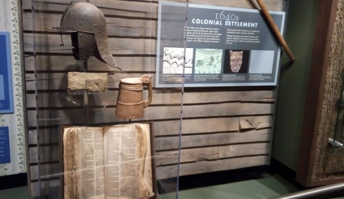 Display case featuring a 1640s helmet, wooden mug, and an open book, showcasing colonial settlement artifacts.
