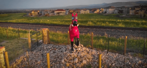 A large, colorful horse statue stands on a rocky mound near a fence, with houses and fields in the background.