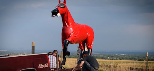 A large red horse statue stands on a field while two people work on it near a pickup truck.