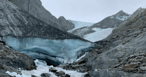 A rugged landscape featuring a glacier with blue ice, rocky terrain, and steep mountain slopes under a cloudy sky.