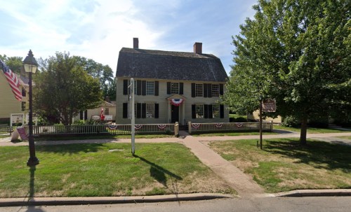 Historic house with green siding, decorative bunting, and a large lawn, surrounded by trees and a lamp post.