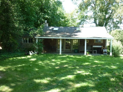 A cozy stone cottage with a porch, surrounded by lush greenery and a grassy yard.