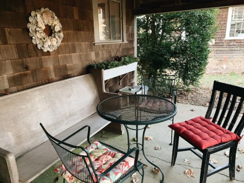 A cozy porch with a wooden bench, a round table, and chairs, surrounded by greenery and a decorative wreath.