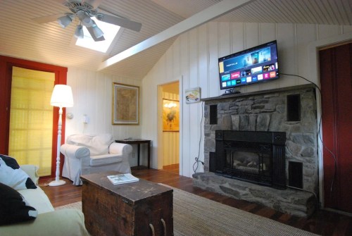 Cozy living room with a stone fireplace, TV on the wall, white furniture, and wooden floors. Bright and inviting atmosphere.