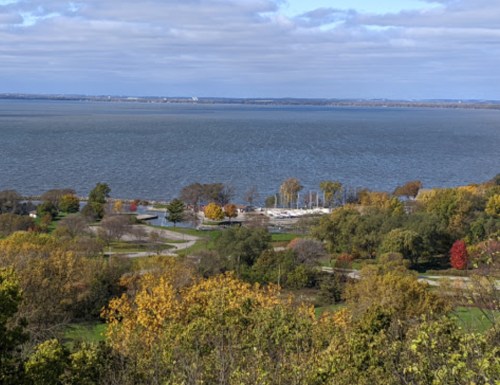 A scenic view of a lake surrounded by trees with autumn foliage and a marina in the distance under a cloudy sky.