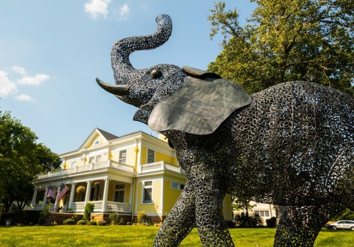 A large metal elephant sculpture stands in front of a yellow house with a porch and American flags.