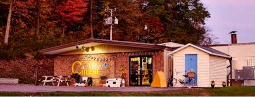 A cozy cheese shop with a brick exterior, surrounded by autumn trees, featuring outdoor seating and decorative elements.