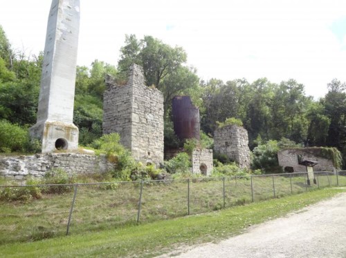Ruins of stone structures surrounded by greenery, with a gravel path and a fence in the foreground.