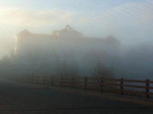 A foggy scene with a building partially obscured by mist, alongside a wooden fence and a cloudy sky.