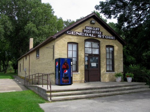 A brick building with a sign reading "High Cliff Post Office General Store," featuring a Pepsi vending machine outside.