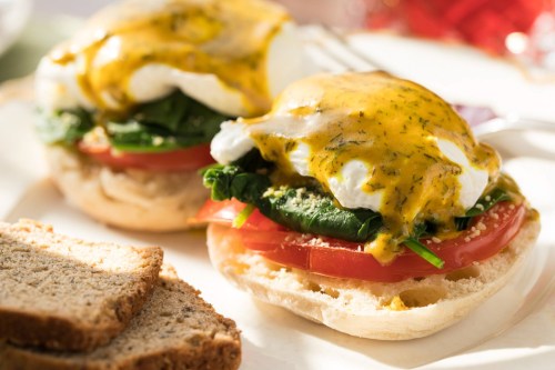 Two poached eggs with hollandaise sauce on a toasted English muffin, layered with spinach and tomato, served with bread.