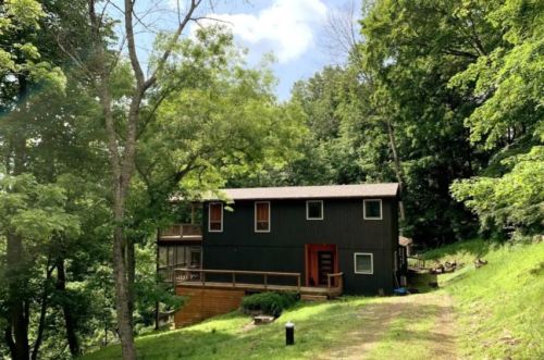 A modern two-story house nestled among lush green trees on a hillside, with a wooden deck and a gravel path.