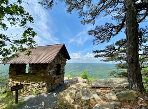 A stone cabin with a metal roof overlooks a lush green valley under a blue sky with scattered clouds.