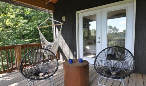 A cozy porch with two black chairs, a round table, and a hammock, surrounded by greenery.
