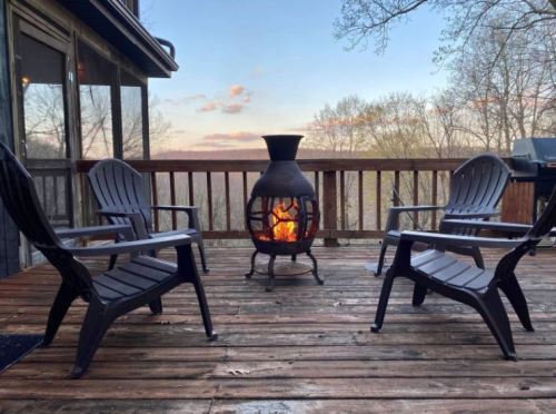 A cozy outdoor deck with four black chairs surrounding a chiminea, glowing with fire against a sunset backdrop.