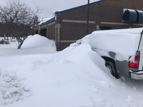 A truck partially buried in deep snow, with snowdrifts against a building in a winter landscape.