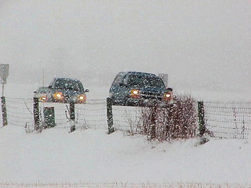 Two cars driving on a snowy road, with heavy snowfall and a fence in the foreground. Visibility is low.