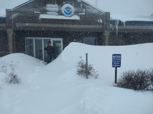 A person stands by a snow-covered entrance, with deep snow drifts and a "Notice" sign visible in the foreground.