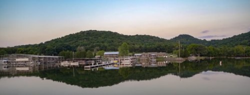 Serene lake view with boats, docks, and lush green hills reflecting in calm water at sunset.