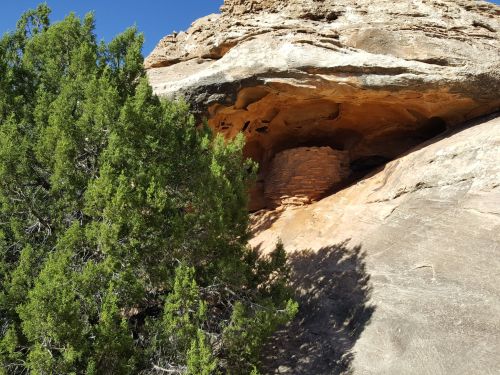 A rocky cliff with a small cave and a stone structure, surrounded by green shrubs under a clear blue sky.