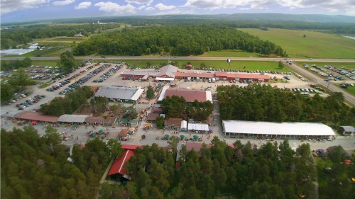Aerial view of a large outdoor event with numerous tents, vehicles, and green trees in the background.