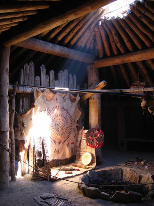 Interior of a traditional dwelling with wooden beams, a circular design on the wall, and a central fire pit.