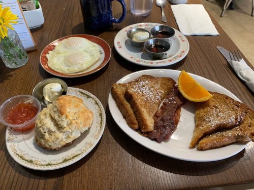 A plate of French toast with orange slice, a biscuit, and a side of egg, with small bowls of syrup and jam.