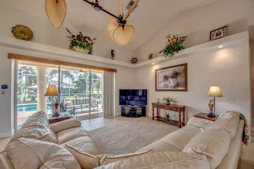 Bright living room with a cream sectional sofa, TV, and large windows overlooking a pool area, adorned with plants.