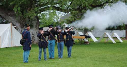 Civil War reenactors firing muskets in a grassy field, with tents and trees in the background. Smoke billows from the guns.