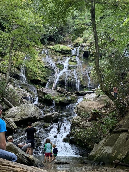 A serene waterfall cascades over rocks, with people enjoying the natural scenery in a lush, green forest.