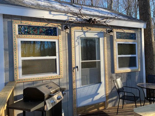 A small cabin with decorative window frames, a door, and a grill on a wooden deck surrounded by trees.