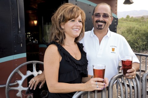 A smiling couple holds drinks outside a vintage train, with scenic greenery in the background.