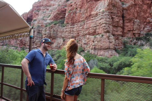 A man and woman stand on a balcony overlooking a rocky landscape, with greenery below and a scenic backdrop.