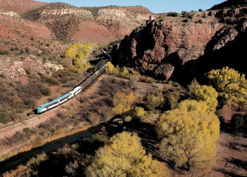 A train travels along a winding track through a rocky landscape with autumn-colored trees and a river below.