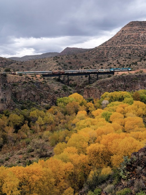 A train crosses a bridge over a canyon, surrounded by vibrant autumn foliage and rugged mountains under a cloudy sky.