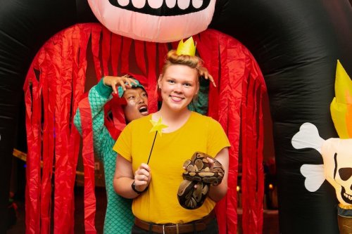 A person in a yellow shirt holds a wand, smiling, while another in a dragon costume playfully poses behind them.