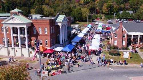 Aerial view of a bustling outdoor market with colorful tents and crowds near historic buildings on a sunny day.