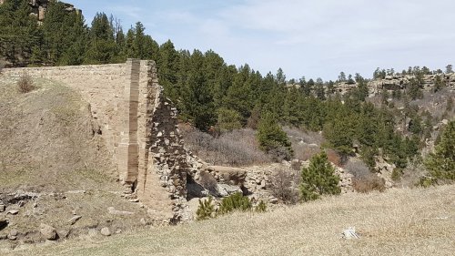 Ruins of a stone structure surrounded by trees and hills under a clear sky.