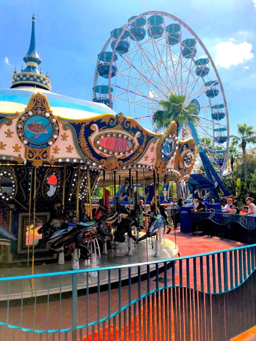 A colorful carousel with ornate horses in the foreground, and a large Ferris wheel in the background under a blue sky.