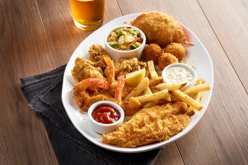 A plate of fried seafood including shrimp, fish, hushpuppies, fries, coleslaw, and dipping sauces, with a beer in the background.