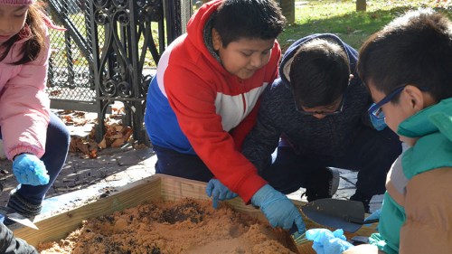 Children wearing gloves play in a sand pit, exploring and digging with tools on a sunny day.