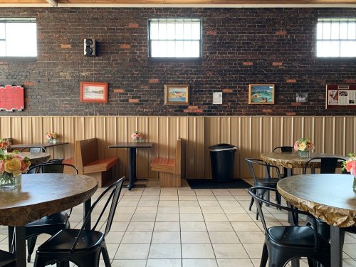 Cozy restaurant interior with wooden tables, black chairs, and floral centerpieces against a brick wall.