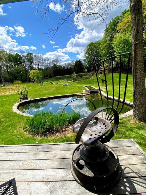 A rustic chair overlooks a serene pond surrounded by lush greenery and a bright blue sky with fluffy clouds.