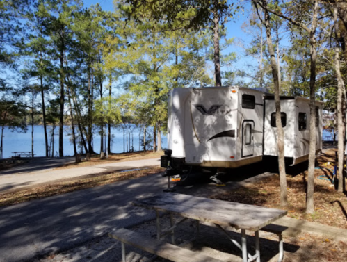 A white RV parked near a lake, surrounded by trees, with a picnic table in the foreground.