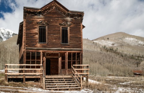 A rustic wooden building with a steep roof, surrounded by snow-covered mountains and bare trees under a cloudy sky.